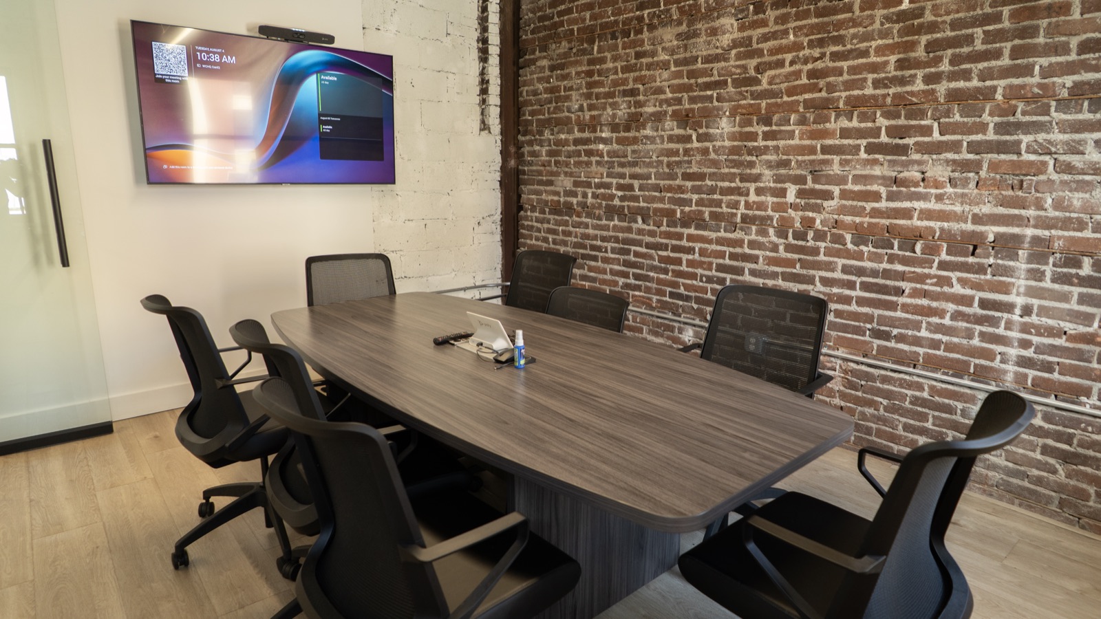 The conference room at The Node: a table for eight against exposed brick, with a wall-mounted display